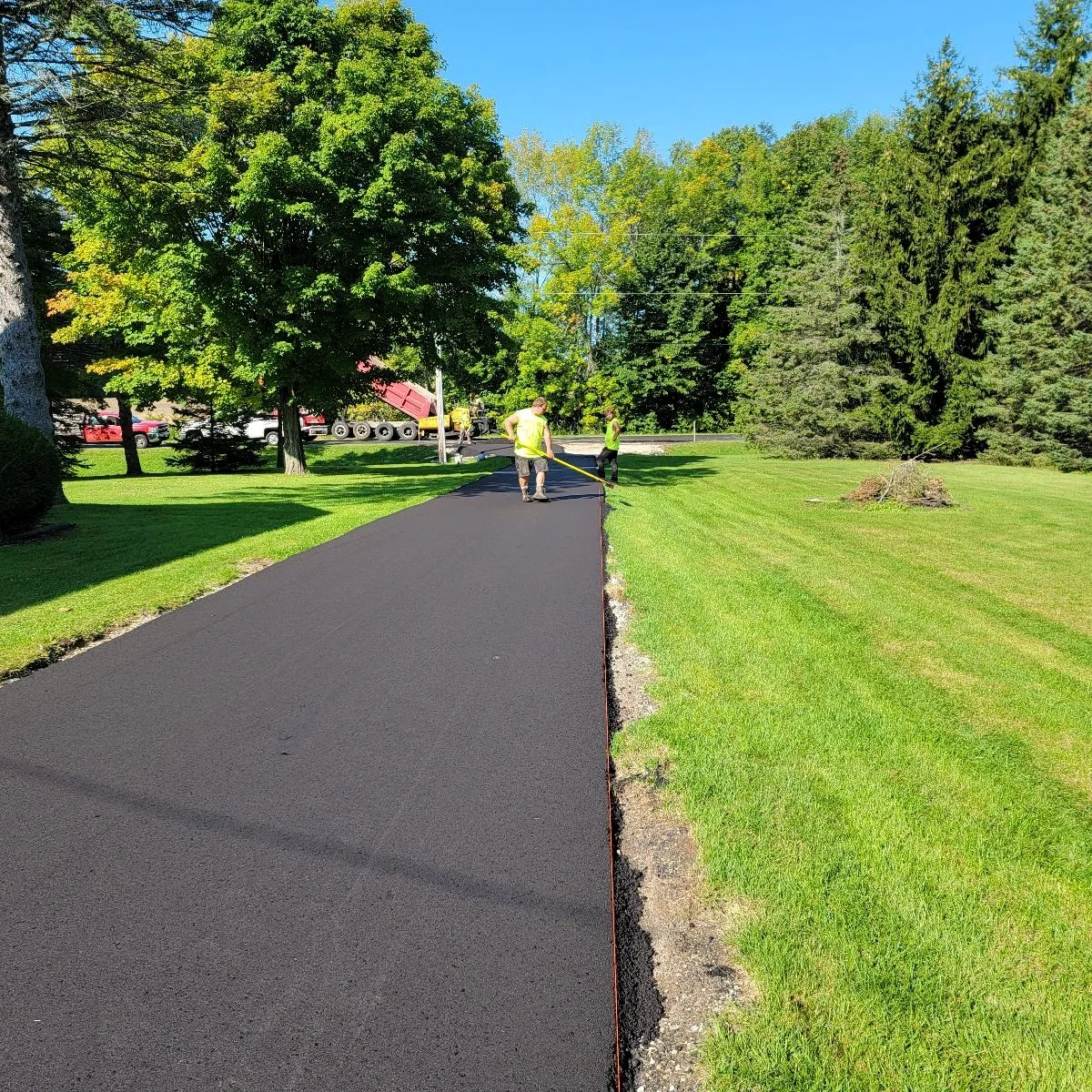 Paving installing a newly paved residential asphalt driveway bordered by lawn and trees.