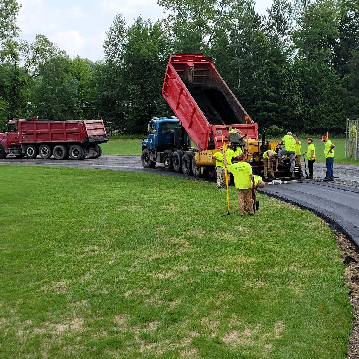 Asphalt paving crew placing fresh pavement on a private road with dump trucks and a paving machine.