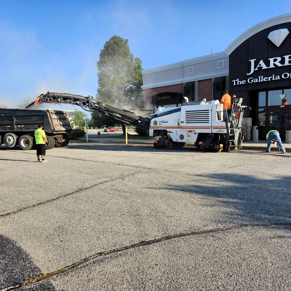 Commercial parking lot milling crew removing deteriorated asphalt with a milling machine and dump truck.
