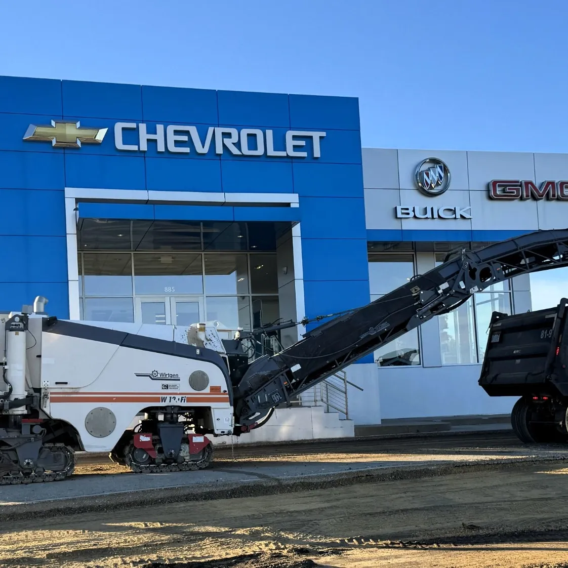 Asphalt milling machine removing pavement at a commercial auto dealership with material loaded into a dump truck.