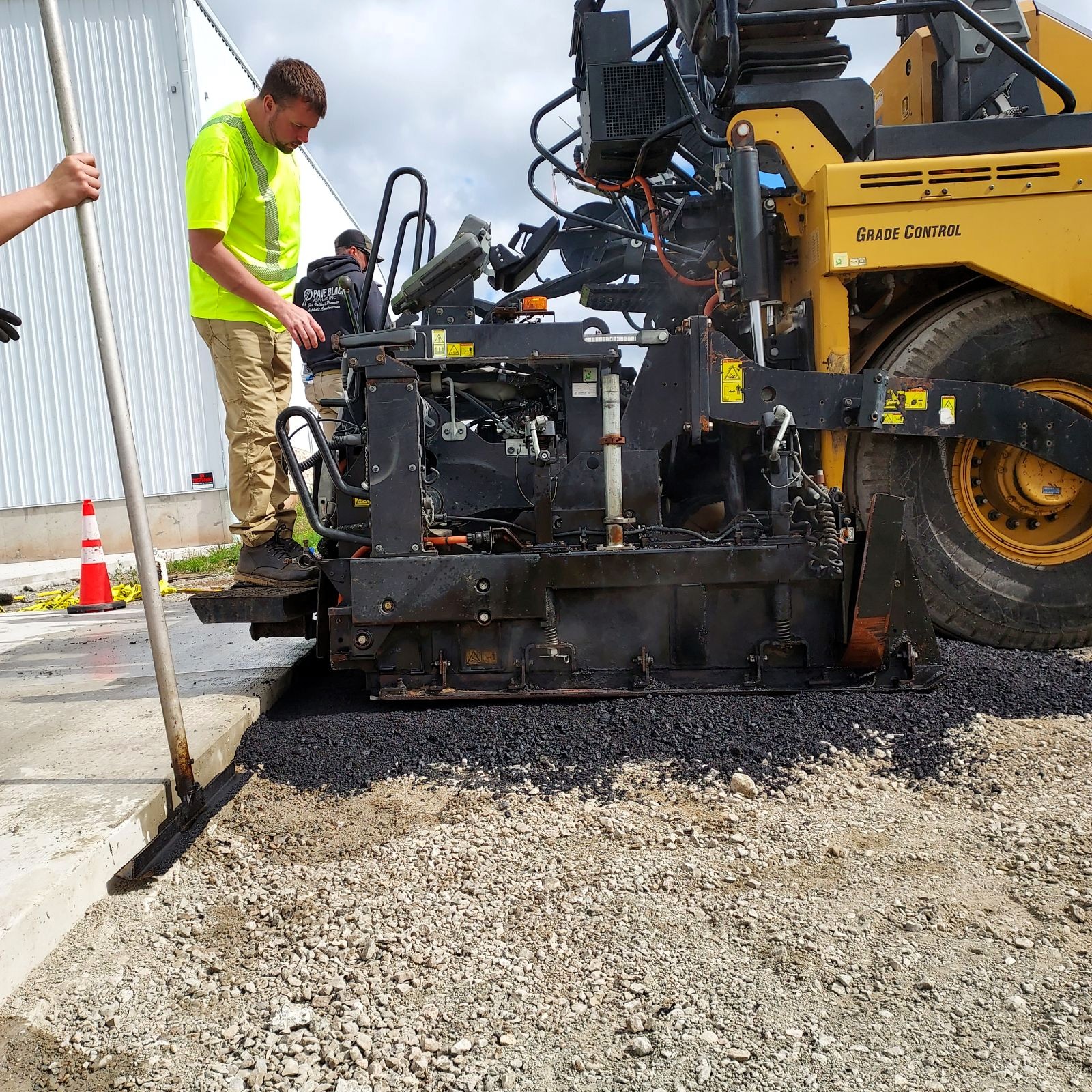 Commercial asphalt paving crew operating a paving machine beside an industrial building.