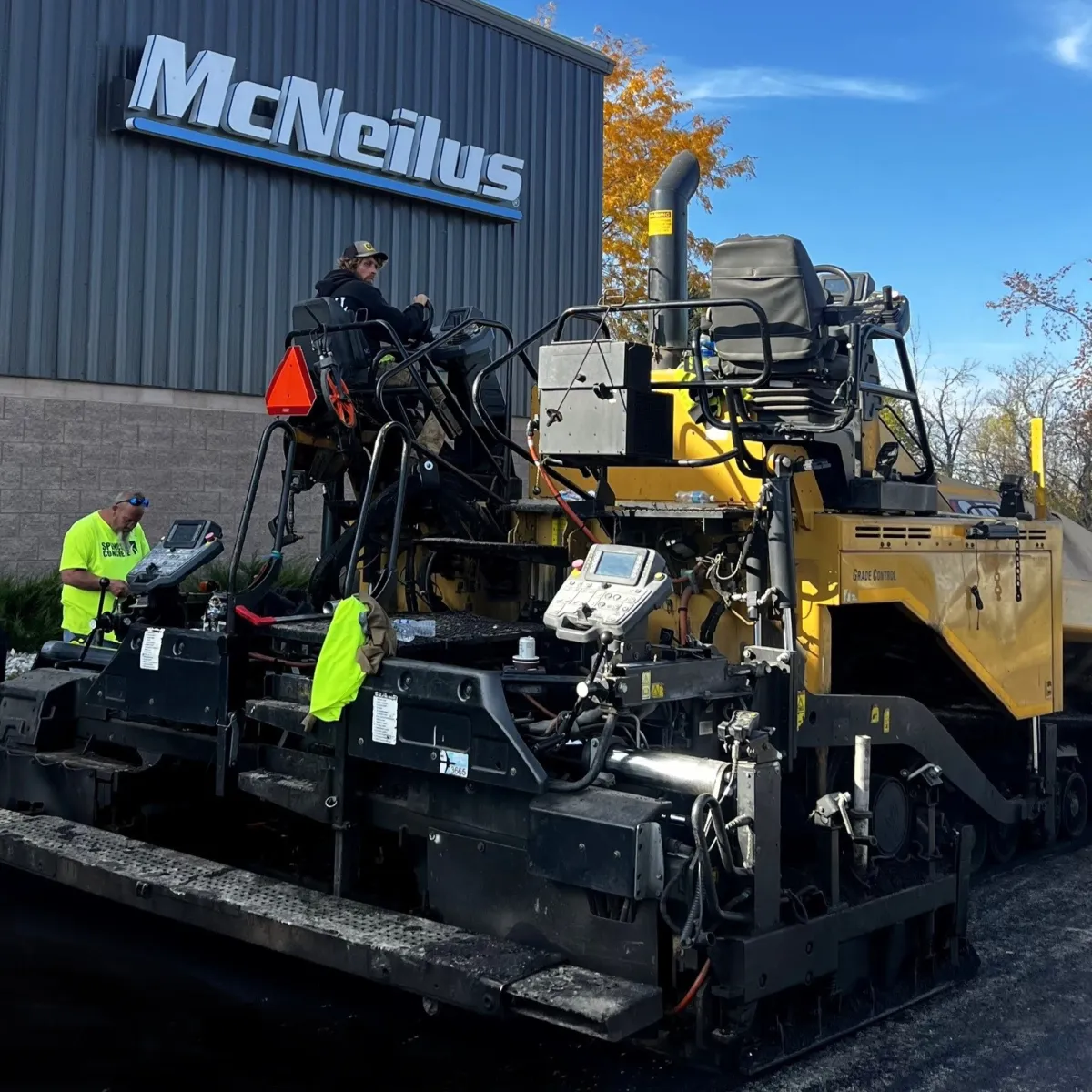 Commercial asphalt paving crew operating a paving machine beside an industrial building.