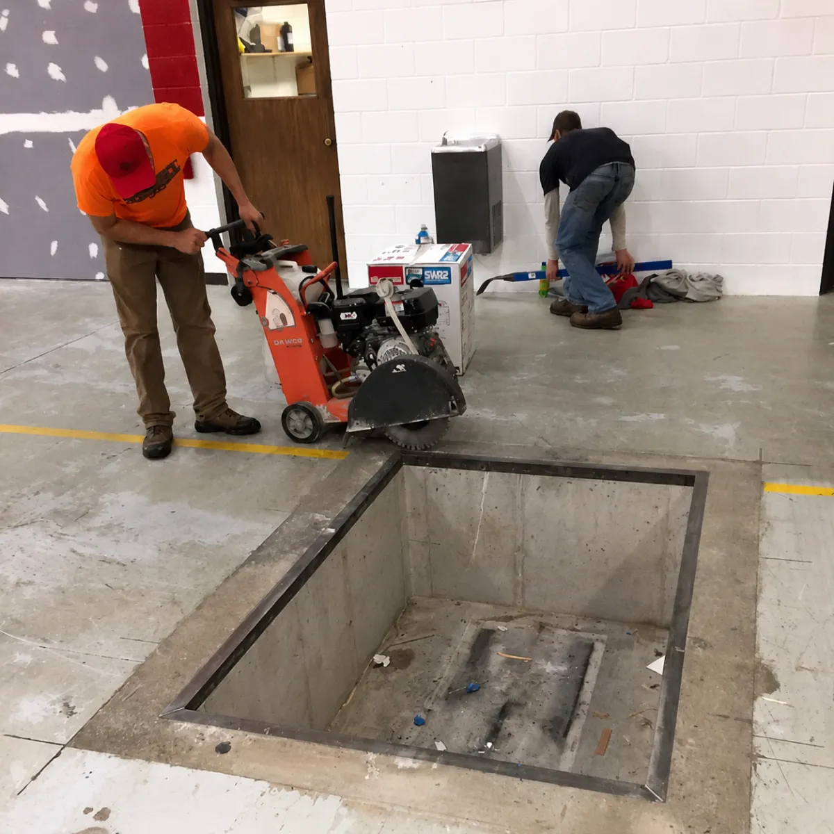 Workers performing indoor concrete saw cutting around a floor opening with a walk-behind concrete saw.