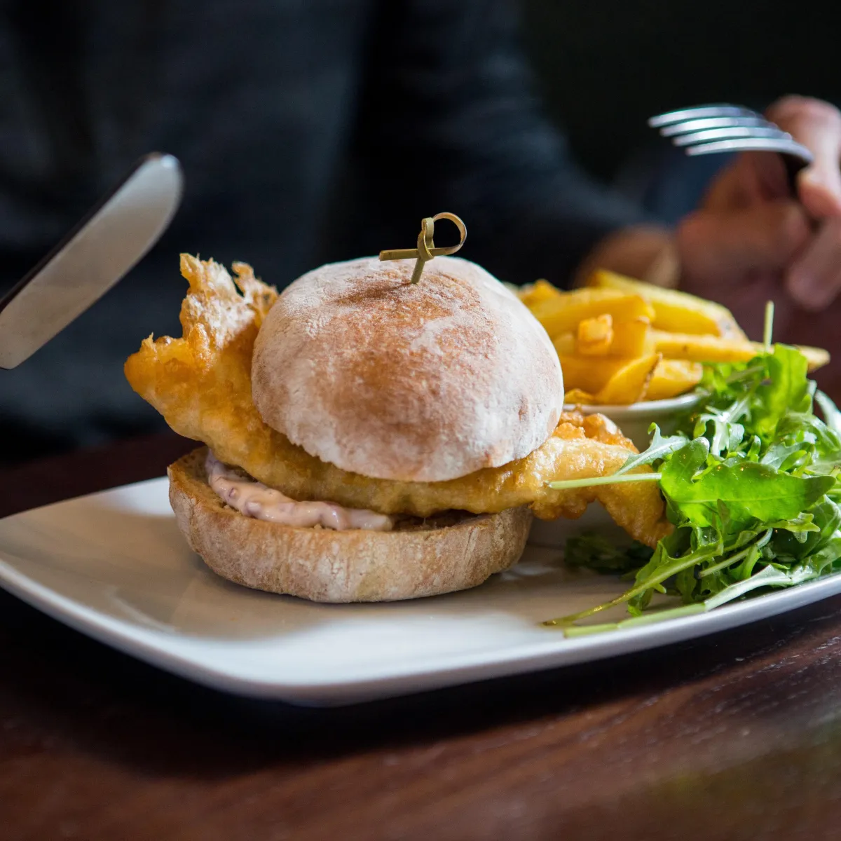 Restaurant photography in Bedfordshire close up of a battered fish in a bun and chips in the background  with a knife about to cut into it