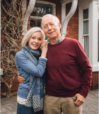 A cheerful middle-aged couple sits at a kitchen table, reviewing documents and smiling as they receive a phone call. Sunlight streams through a window, and a laptop is open in front of them. The scene conveys relief and optimism after a successful property sale.
