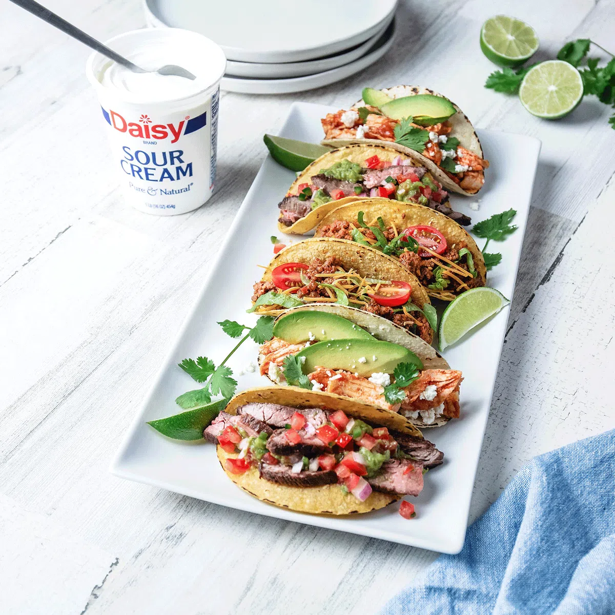Colorful taco platter featuring steak and chicken tacos with avocado, cilantro, and fresh garnishes, styled with lime wedges and Daisy sour cream.