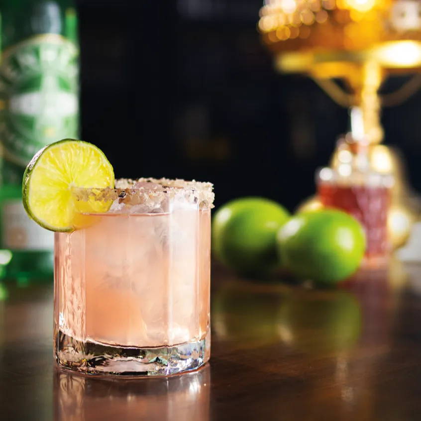 Citrus gin cocktail with lime garnish and salted rim, photographed on a bar with fresh limes and ambient lighting for a cozy nightlife setting.