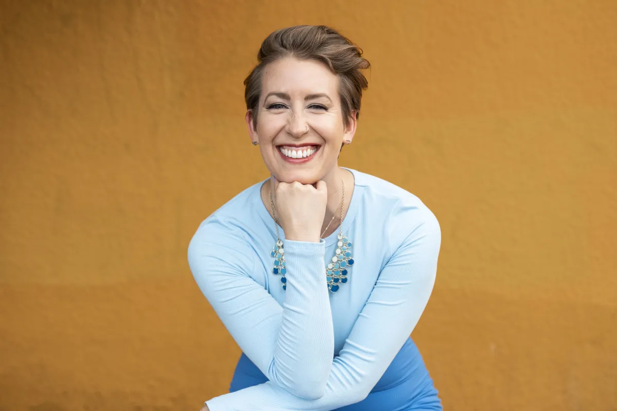 woman with yellow background, short hair, blue shirt, big smile, fist under chin looking confident