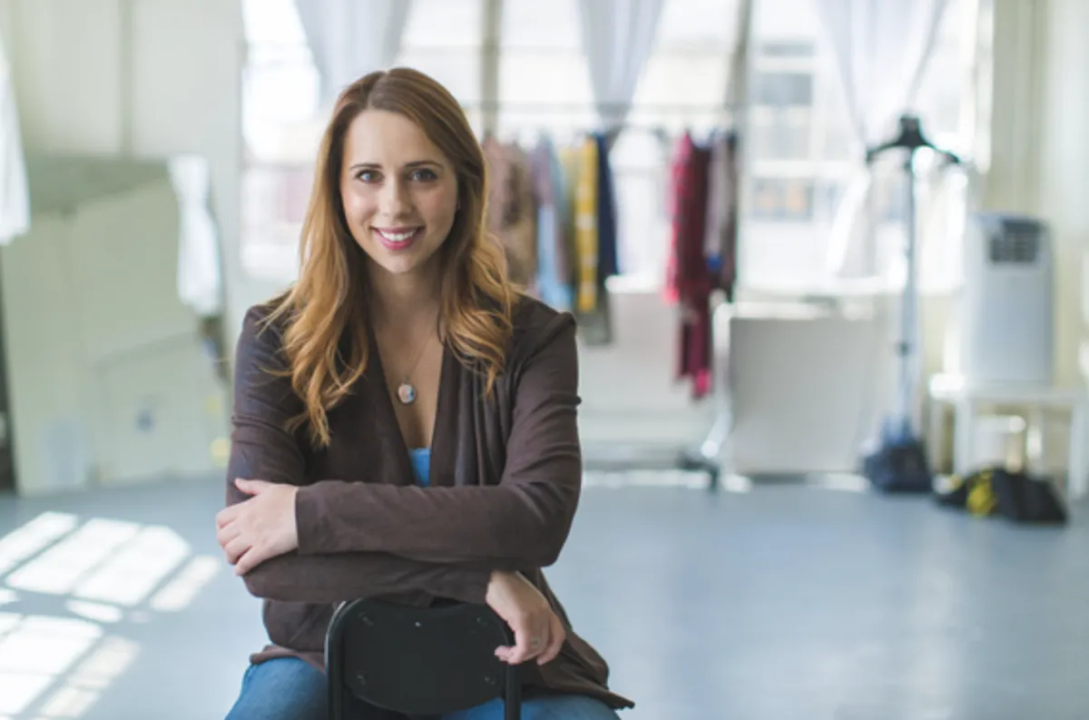 woman sitting backwards on a chair wearing brown jacket and smilng, arms crossed