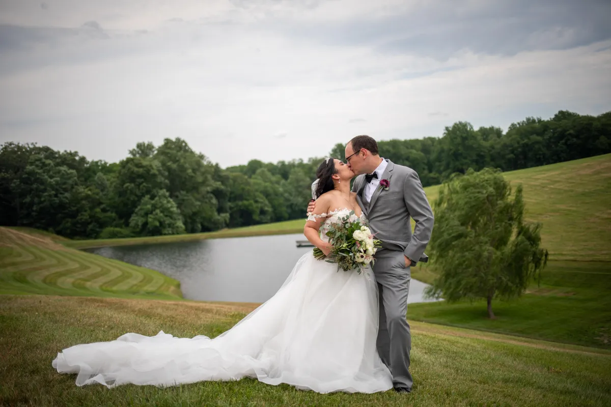 Bride and groom sharing a kiss on the hilltop at Morningside Inn, with a scenic pond, rolling hills, and lush trees in the background