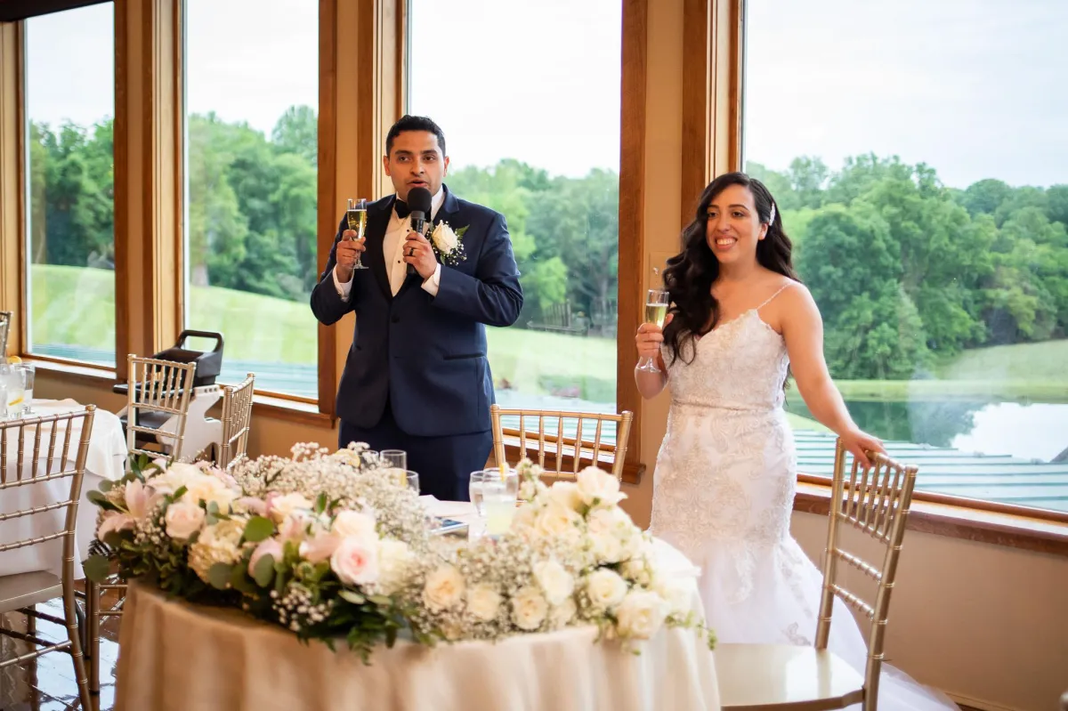 Bride and groom giving a wedding toast at Morningside Inn, standing behind a floral-covered sweetheart table with scenic pond views through large windows