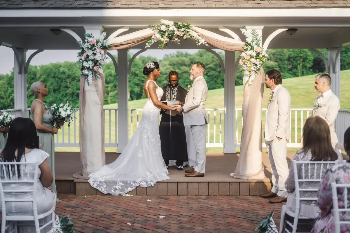 Bride and groom exchanging vows under a flower-adorned gazebo at Morningside Inn, surrounded by their wedding party and guests on a sunny day