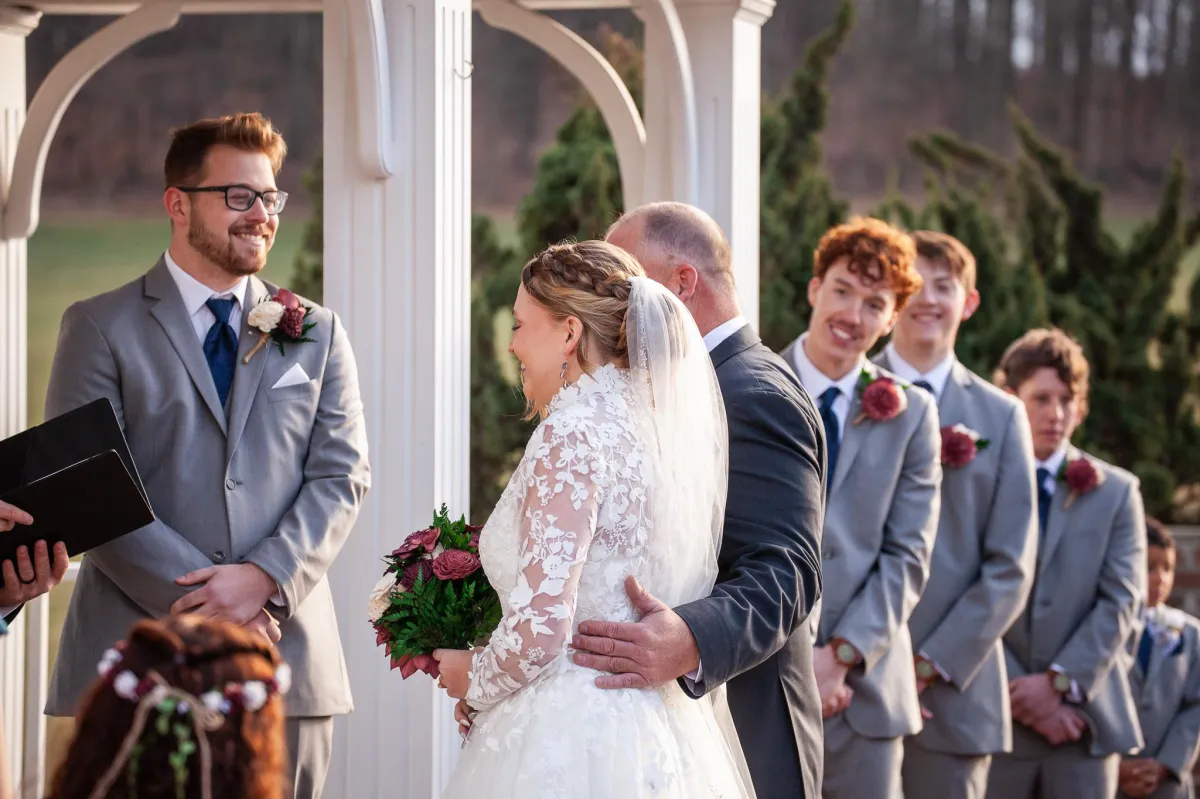 Bride smiling as she walks down the aisle with her father at Morningside Inn, greeted by the groom and groomsmen in gray suits