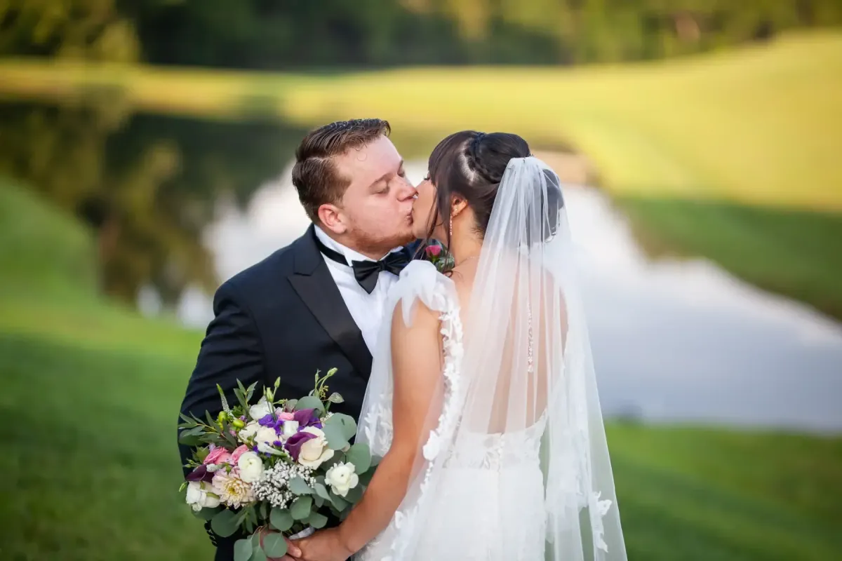 Bride and groom share a kiss by the pond at Morningside Inn wedding venue in Frederick, MD, with the bride in a lace gown and veil and the groom in a black tuxedo, holding a bouquet of purple and white flowers.