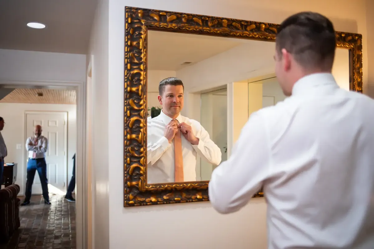 Groom adjusting his tie in front of a large ornate mirror in the groom’s suite at Morningside Inn wedding venue in Frederick, Maryland, with groomsmen in the background.
