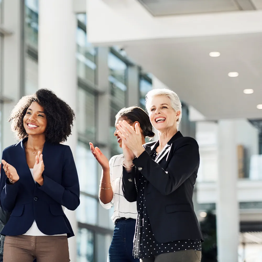 Three professional women in business attire smiling and clapping outdoors, representing confident connection and enthusiastic support for storytelling success.