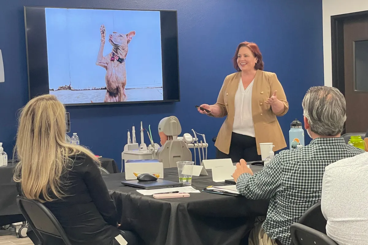 Mary Czarnecki leading a storytelling workshop in front of a seated audience, with a slide featuring a dog and dental tools, illustrating real-world applications of strategic communication.