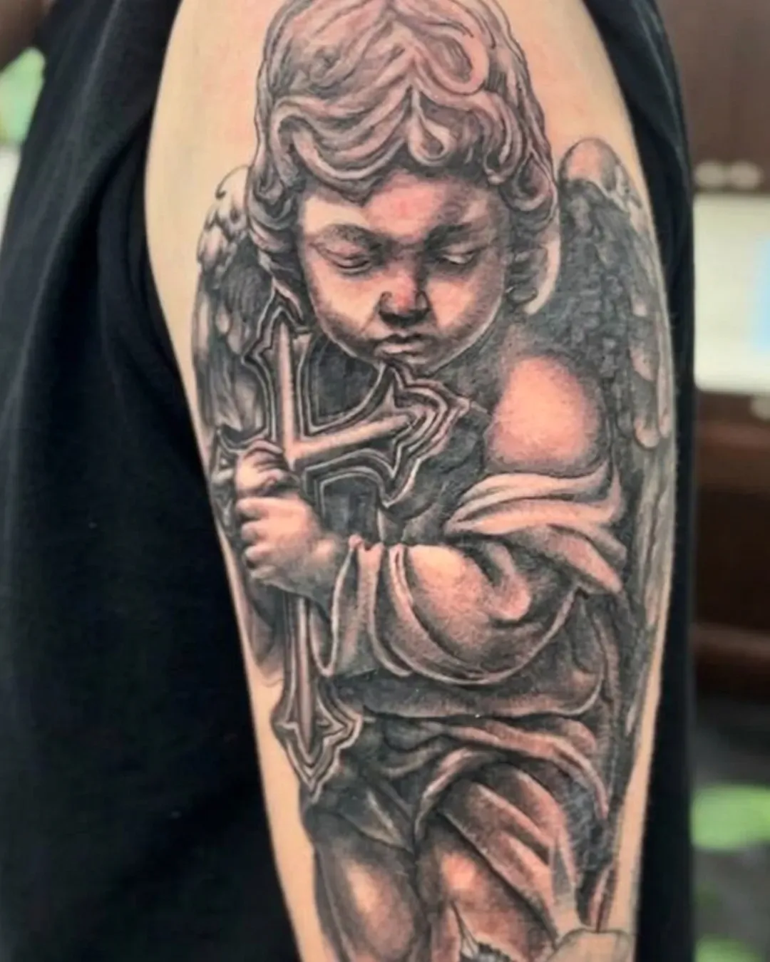  Black and gray tattoo of a baby angel statue, holding a cross on the bicep in Oceanside, California