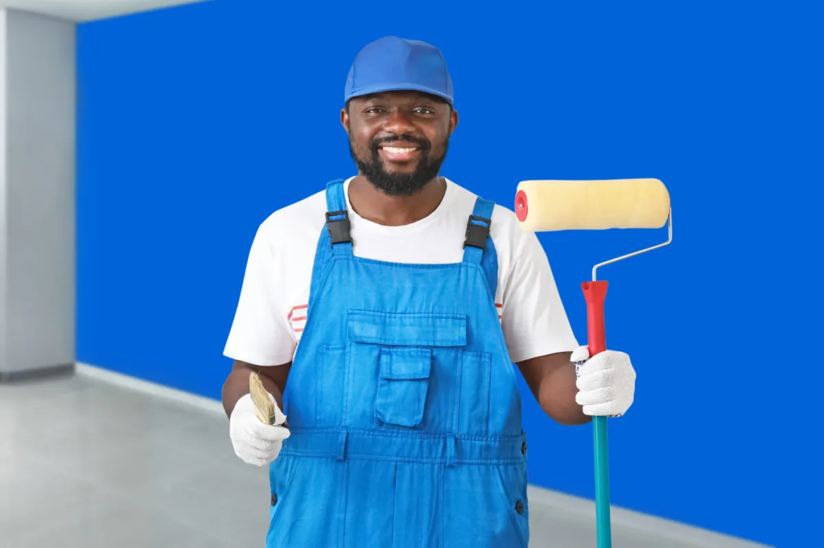 Professional painter in blue overalls holding a roller and brush, standing in front of a freshly painted blue wall