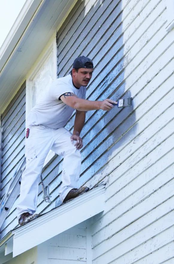 Professional painter applying fresh paint to the exterior siding of a house using a brush, showcasing the transformation of the home's appearance.