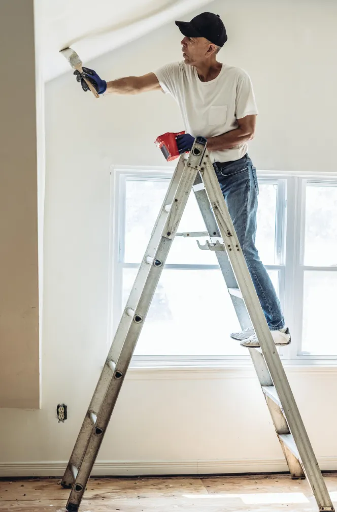 A professional painter standing on a ladder, painting the upper walls of a room with a roller brush during an interior painting project.