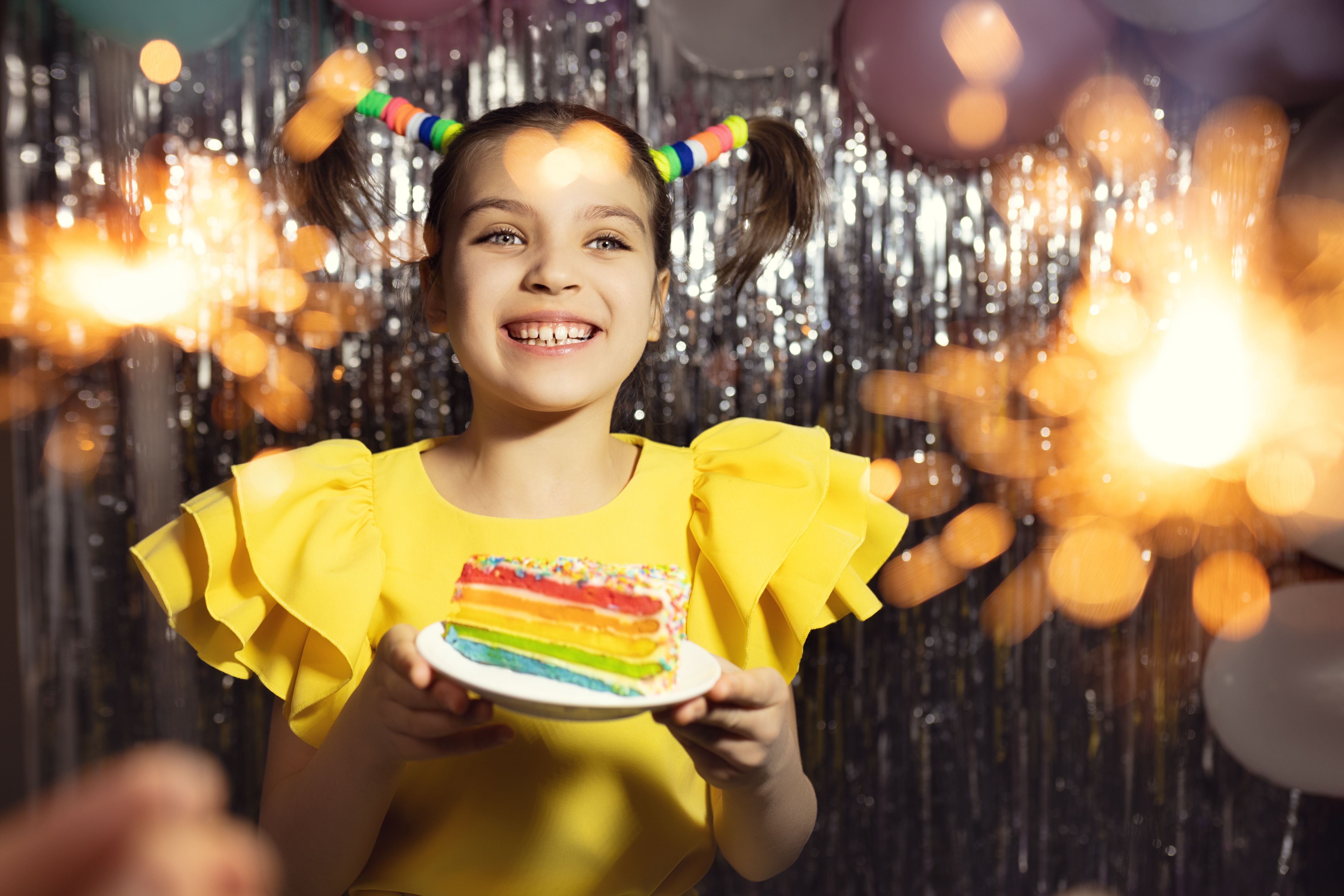 Smiling girl in yellow dress holding colorful rainbow birthday cake, celebrating with festive decorations and sparklers, embodying joy and excitement for a Top Gun birthday party.
