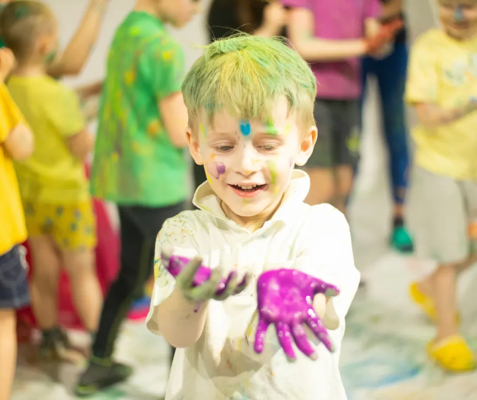 Child with colorful paint on hands and face, smiling joyfully, surrounded by other children in a vibrant, playful environment, representing fun activities at Top Gun Ohio's events for kids.