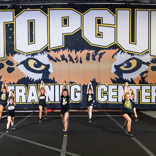 Young cheerleaders performing in front of a large "TOP GUN TRAINING CENTER" banner, showcasing teamwork and athleticism in a gymnastics facility.