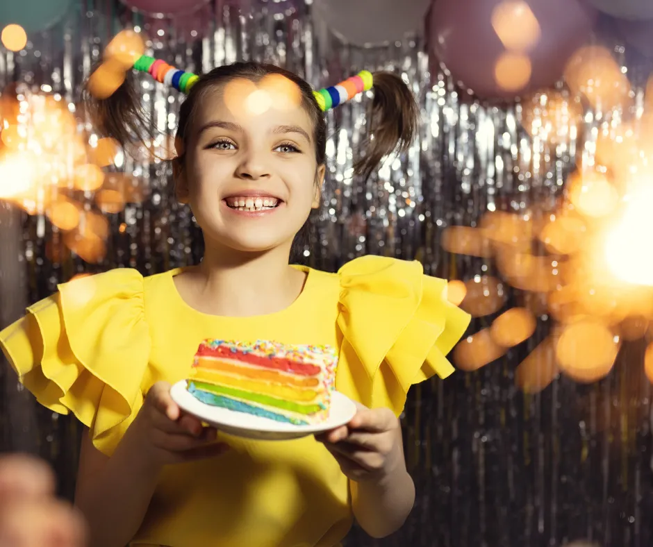 Smiling girl in yellow dress holding a colorful rainbow cake, celebrating at a festive birthday party with sparkles and balloons in the background.