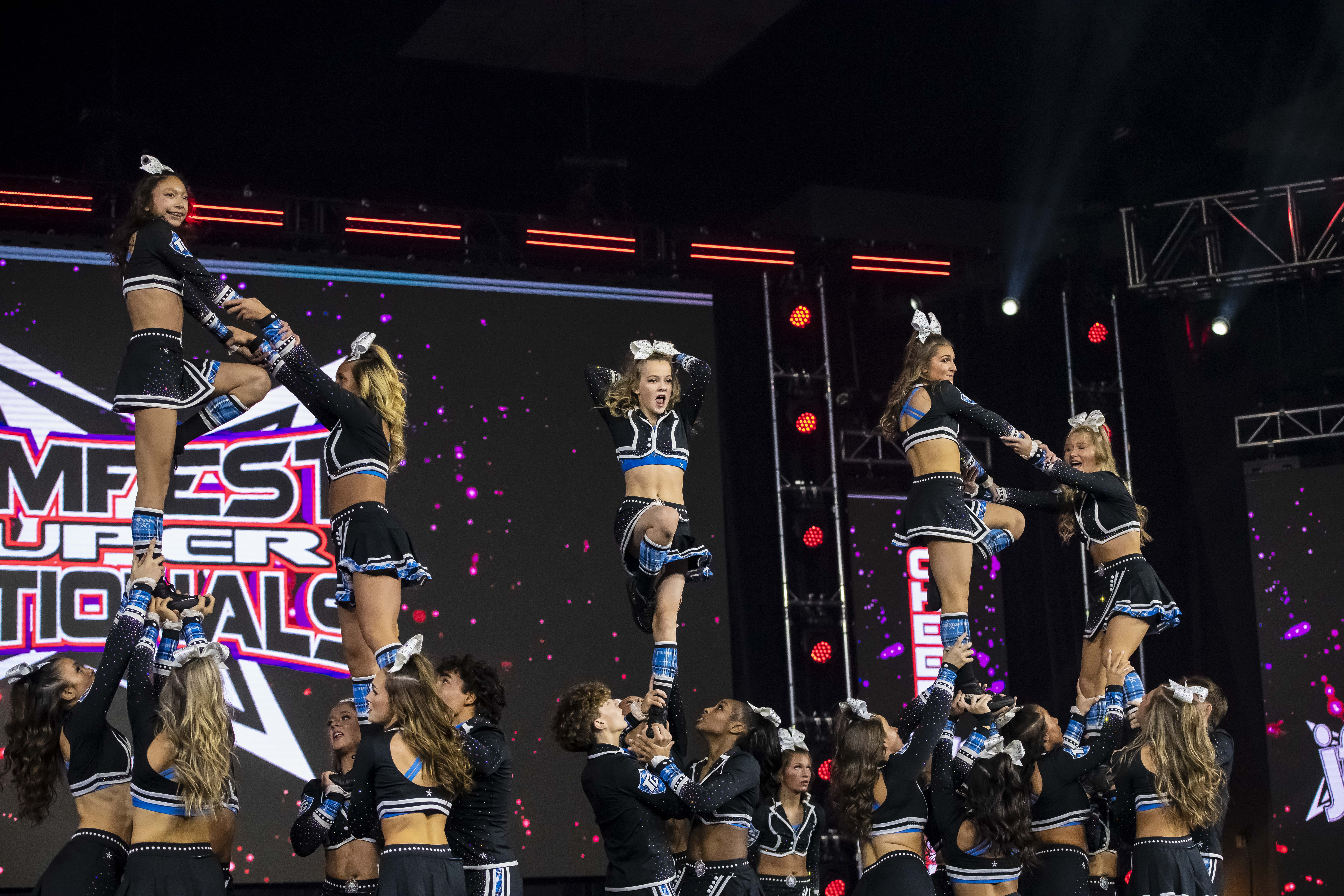 Cheerleaders performing a stunt at a competition, showcasing teamwork and athleticism, with a vibrant backdrop featuring "Jamfest Super Nationals" branding.