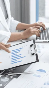 Person analysiert Geschäftsdaten auf einem Laptop, während eine andere Hand ein Klemmbrett mit Balkendiagrammen hält – Fokus auf Datenanalyse und Reporting.