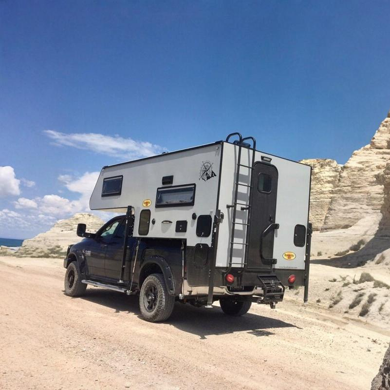 A black truck carrying a white slide-in camper parked on a sandy desert road.