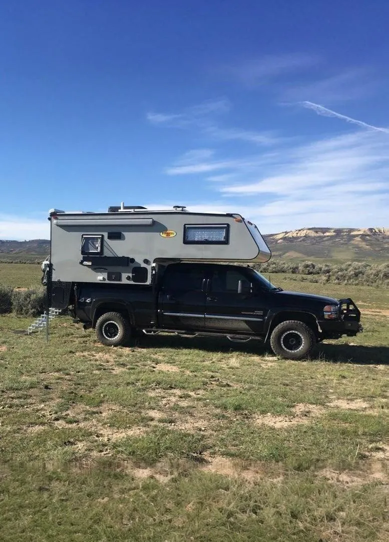 A black truck with a gray slide-in camper parked on a grassy plain under a clear sky.
