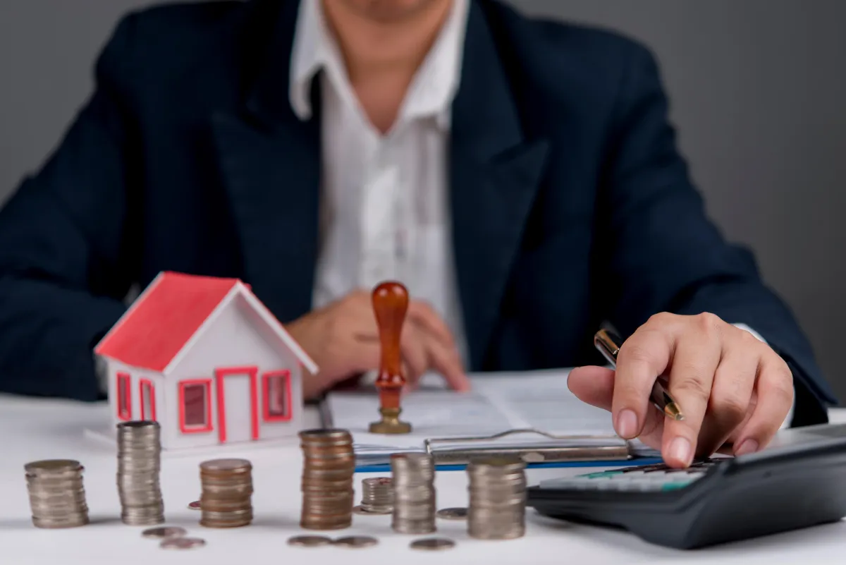 stacks of coins and toy house in background with man and papers