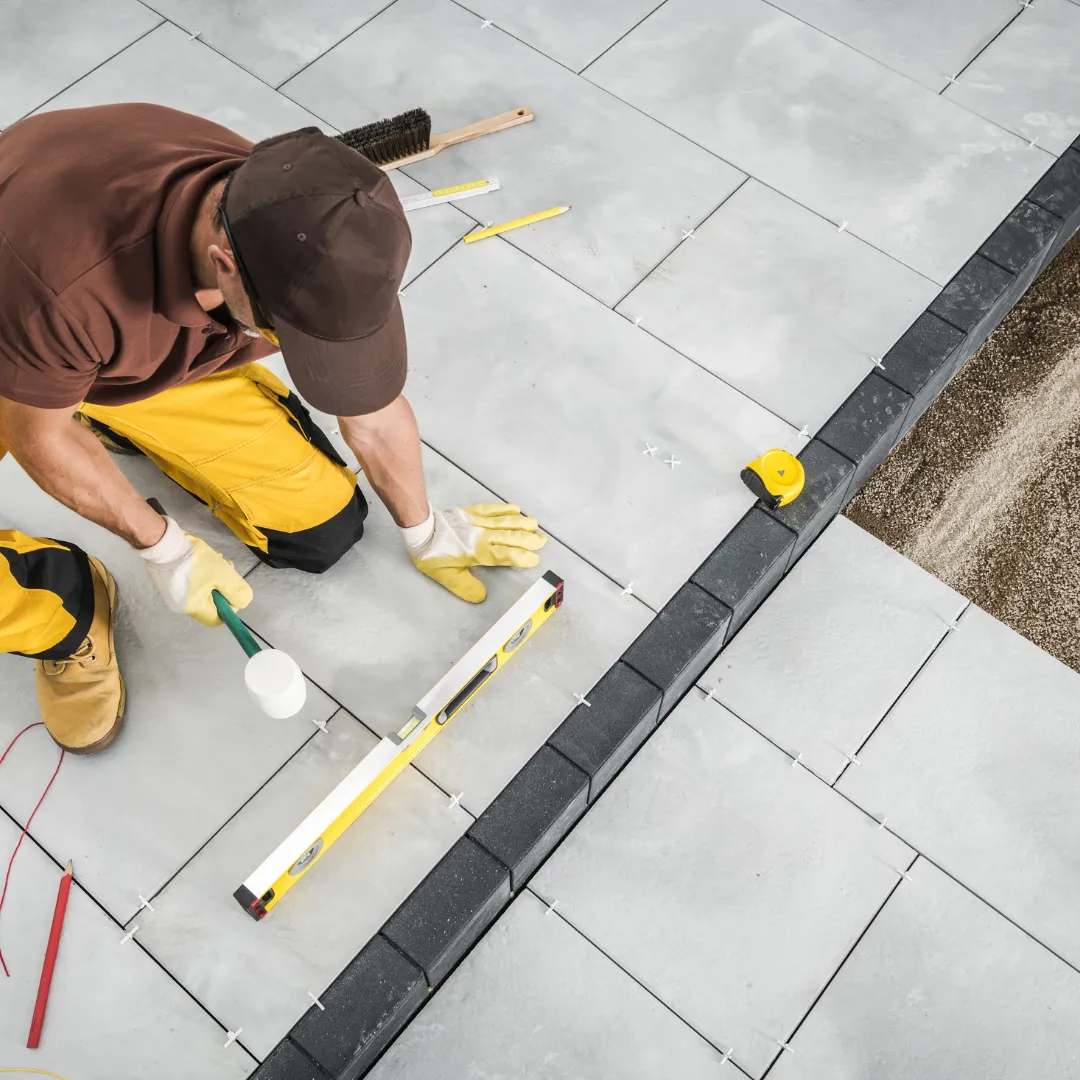 A worker in yellow pants and gloves lays gray paving stones using a level and rubber mallet. Tools like a brush and tape measure are nearby.