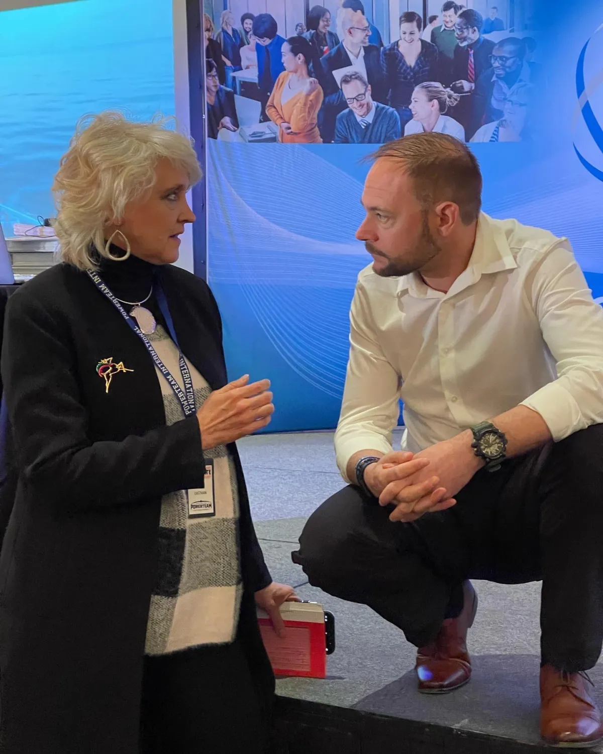 Baz Porter in a white shirt, engaged in conversation with an older woman with white hair, wearing a black coat and a badge, at a professional event. They are standing in front of a backdrop depicting a diverse group of people in a business setting.