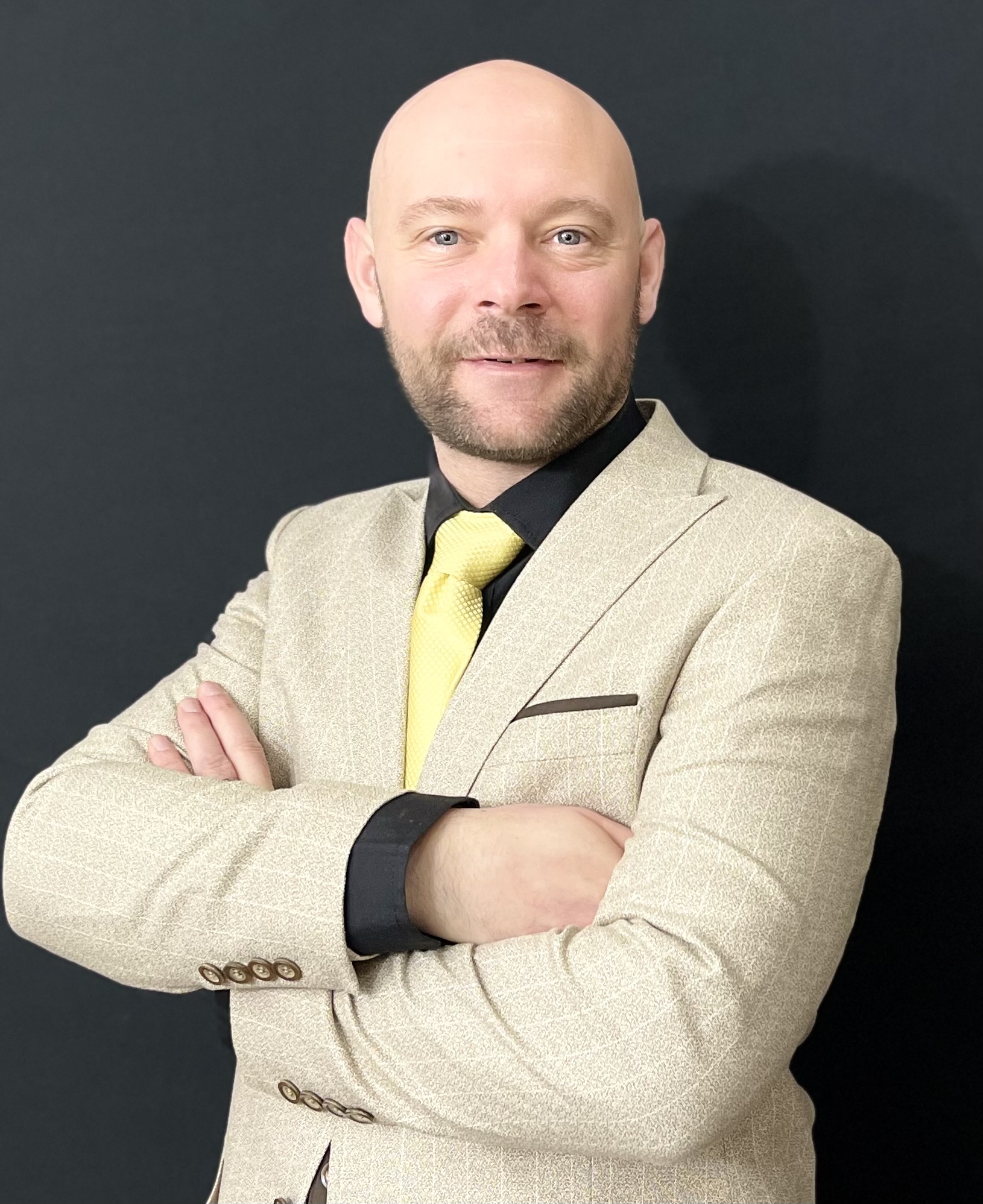 Baz Porter, a transformative leadership coach, wearing a beige suit with a yellow tie and black shirt, standing confidently with arms crossed against a dark background. Symbolizing resilience, empowerment, and success.