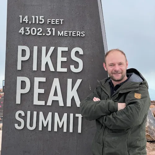 Baz Porter standing confidently at Pikes Peak Summit, marked at 14,115 feet (4,302.31 meters), with a cloudy mountain backdrop. Achievement and resilience symbolized through the iconic landmark and his poised stance.