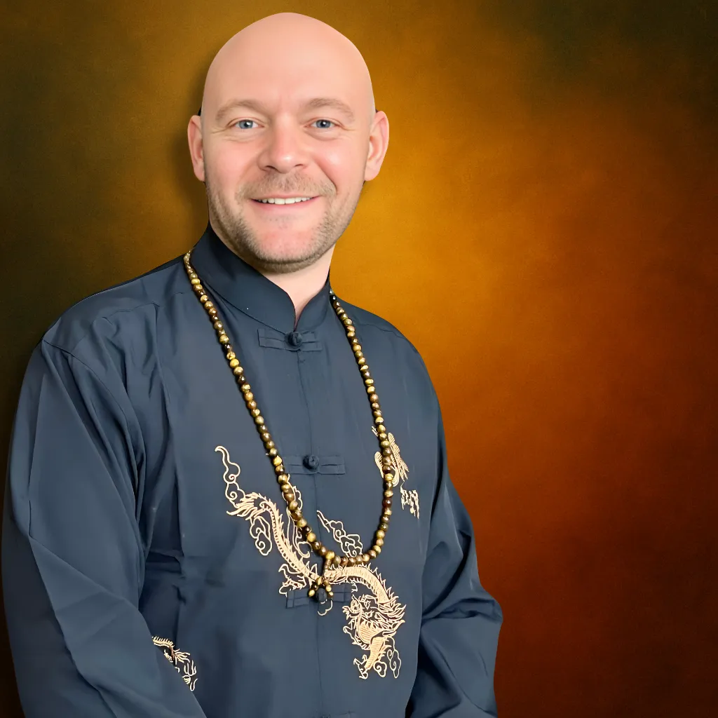 Baz Porter, 2X international bestselling author and transformational coach, wearing a black and gold dragon-embroidered traditional jacket with meditation beads, smiling confidently in front of a radiant orange background.