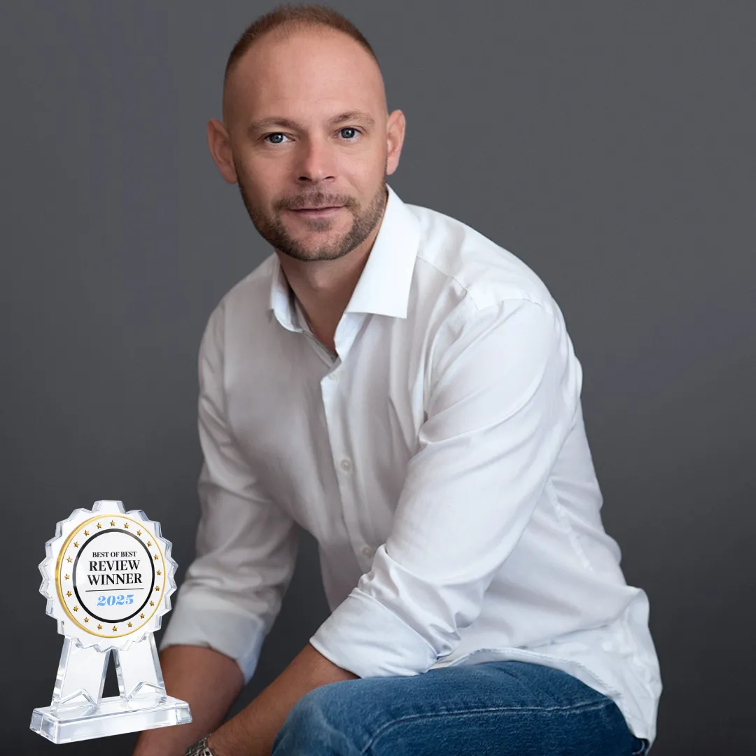 Portrait of Baz Porter, business strategist and founder of Baz Porter LLC™, seated in a studio setting wearing a white shirt and jeans, holding a crystal acrylic “Best of Best Review Winner 2025” award. The premium 3D trophy features a circular medallion with gold star detailing and engraved text, symbolizing excellence, credibility, and industry recognition in executive coaching and leadership development.
