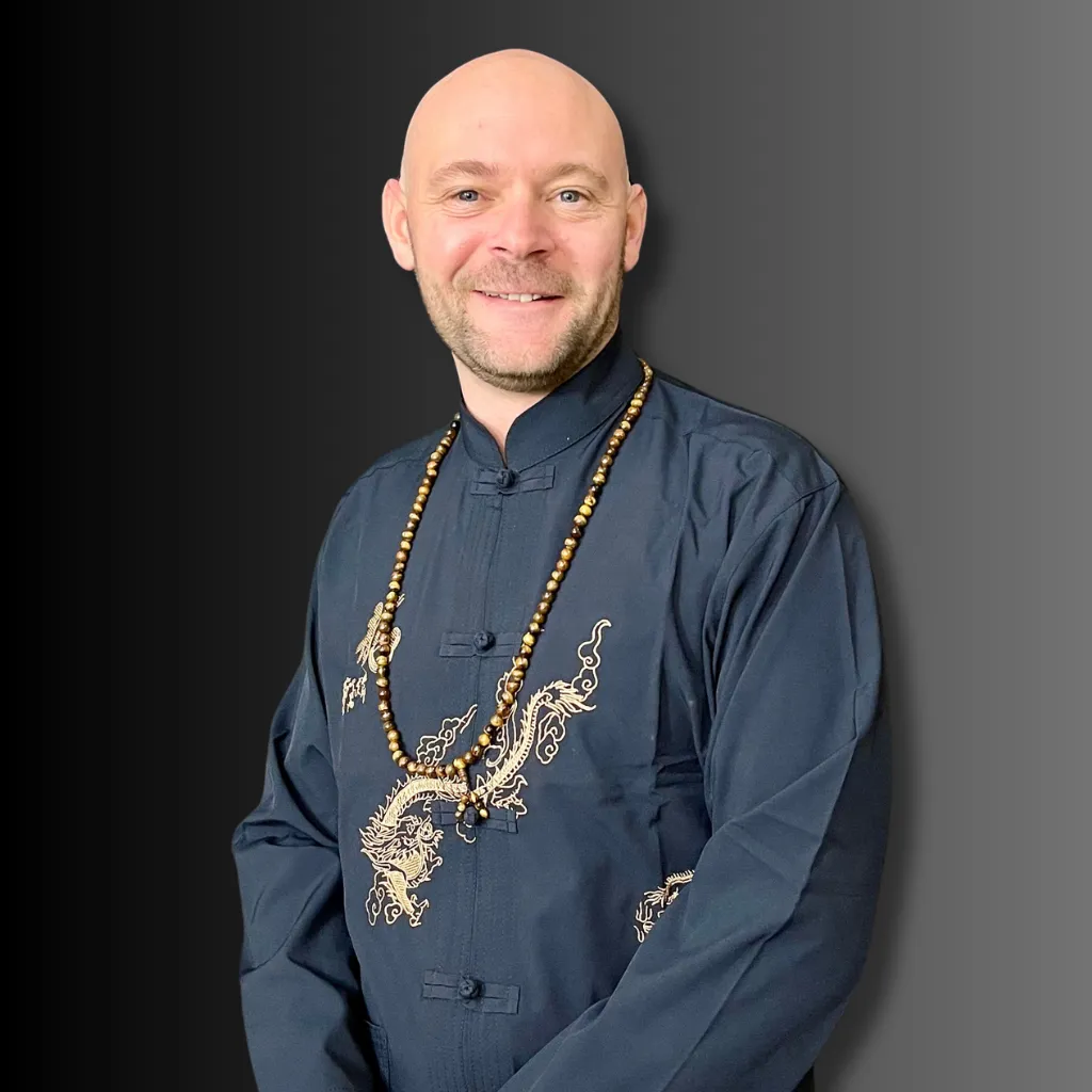 Baz Porter wearing a traditional dark navy outfit with gold dragon embroidery and a beaded necklace, standing confidently against a gradient background.