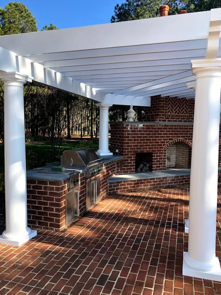 Outdoor kitchen with stainless steel grills and brickwork under a white pergola, surrounded by trees, showcasing high-quality craftsmanship for residential spaces.