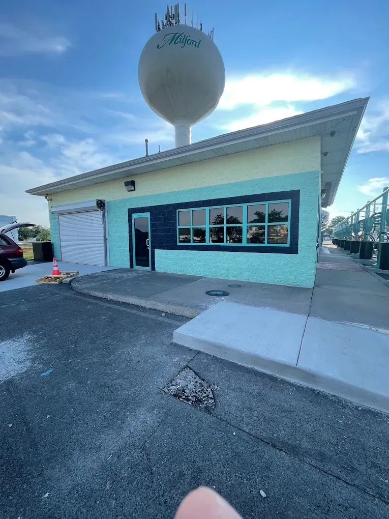 Exterior view of a colorful commercial building with a blue and teal facade, featuring large windows, a roll-up door, and a water tower labeled "Milford" in the background, representing professional painting services.