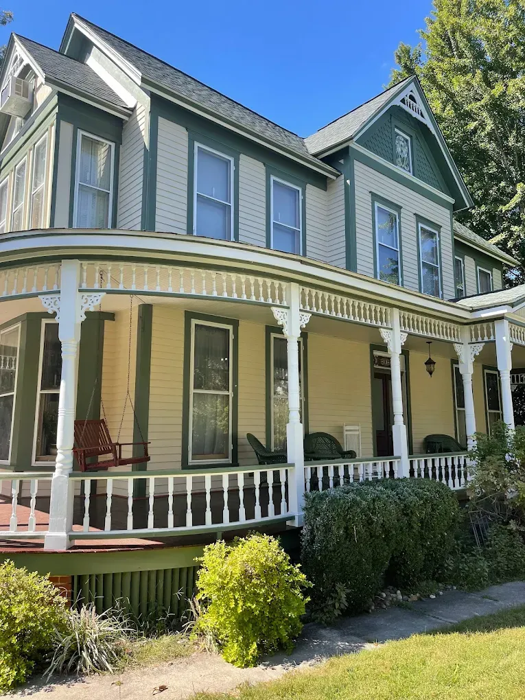 Victorian-style house with green and yellow exterior, featuring a wraparound porch, decorative trim, and lush landscaping, showcasing exterior painting services by Victory Painting LLC.