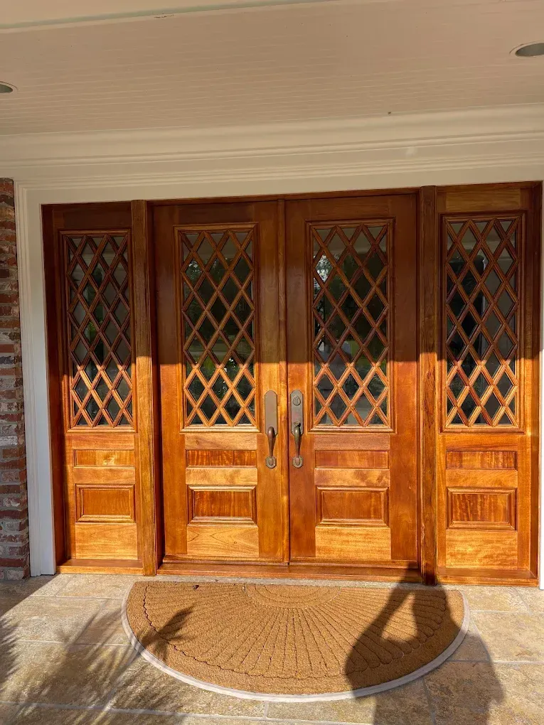 Double wooden front doors with diamond-patterned glass panels and polished finish, leading to a residential entryway, complemented by a round welcome mat.