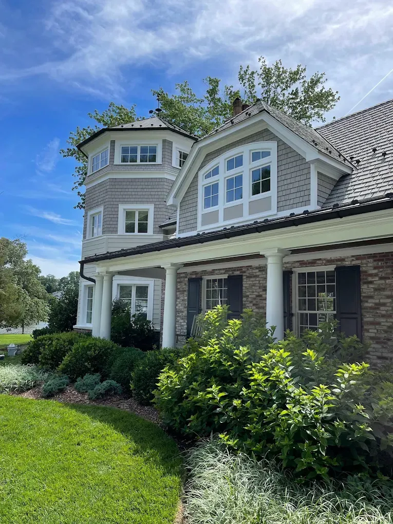 Exterior view of a well-maintained house with a turret, featuring a combination of stone and siding, surrounded by lush greenery and a manicured lawn, illustrating high-quality residential painting and curb appeal.