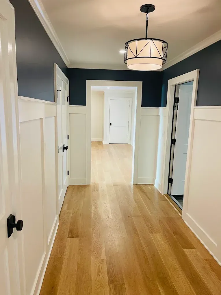 Modern hallway featuring light hardwood flooring, dark blue accent wall, white wainscoting, and a decorative ceiling light fixture, showcasing a freshly painted interior suitable for residential spaces.