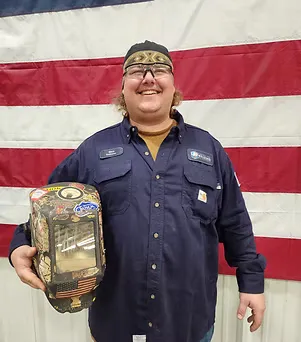 Man in dark uniform holding a decorated welding helmet, standing before a flag.