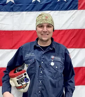 Welder in blue uniform holding a helmet with stickers, standing before a flag.
