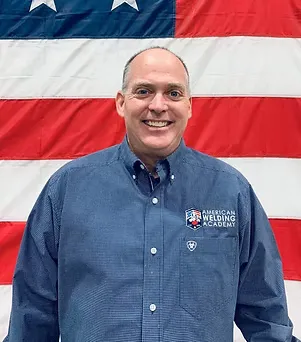Man in blue shirt standing in front of an American flag background.